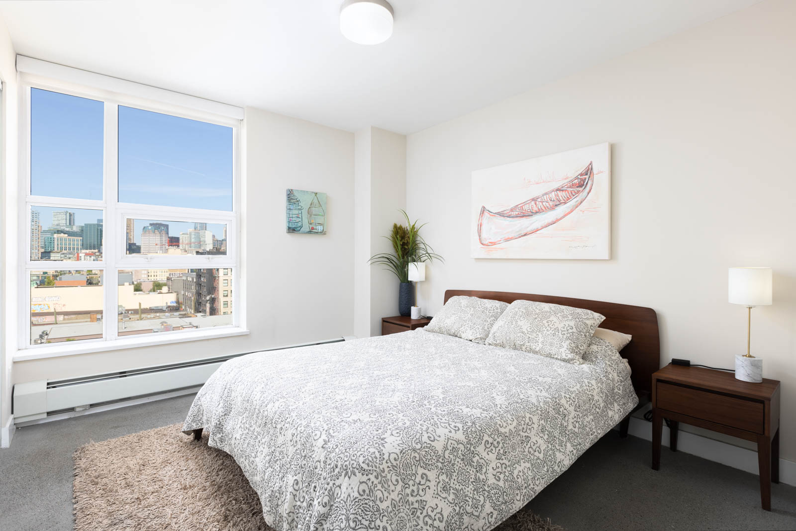 A bedroom with a large window showing a cityscape view, a double bed with a patterned duvet, two side tables with lamps, and wall art above the bed.
