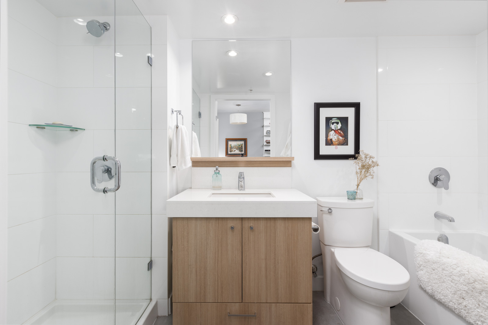 A modern bathroom features a glass shower, wooden vanity with sink, toilet, and white bathtub. A framed picture and a small plant are on the vanity. Walls and tiles are white.