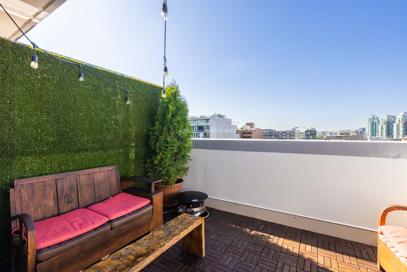 Rooftop patio with wooden seating, pink cushions, a green plant, string lights, and cityscape view.