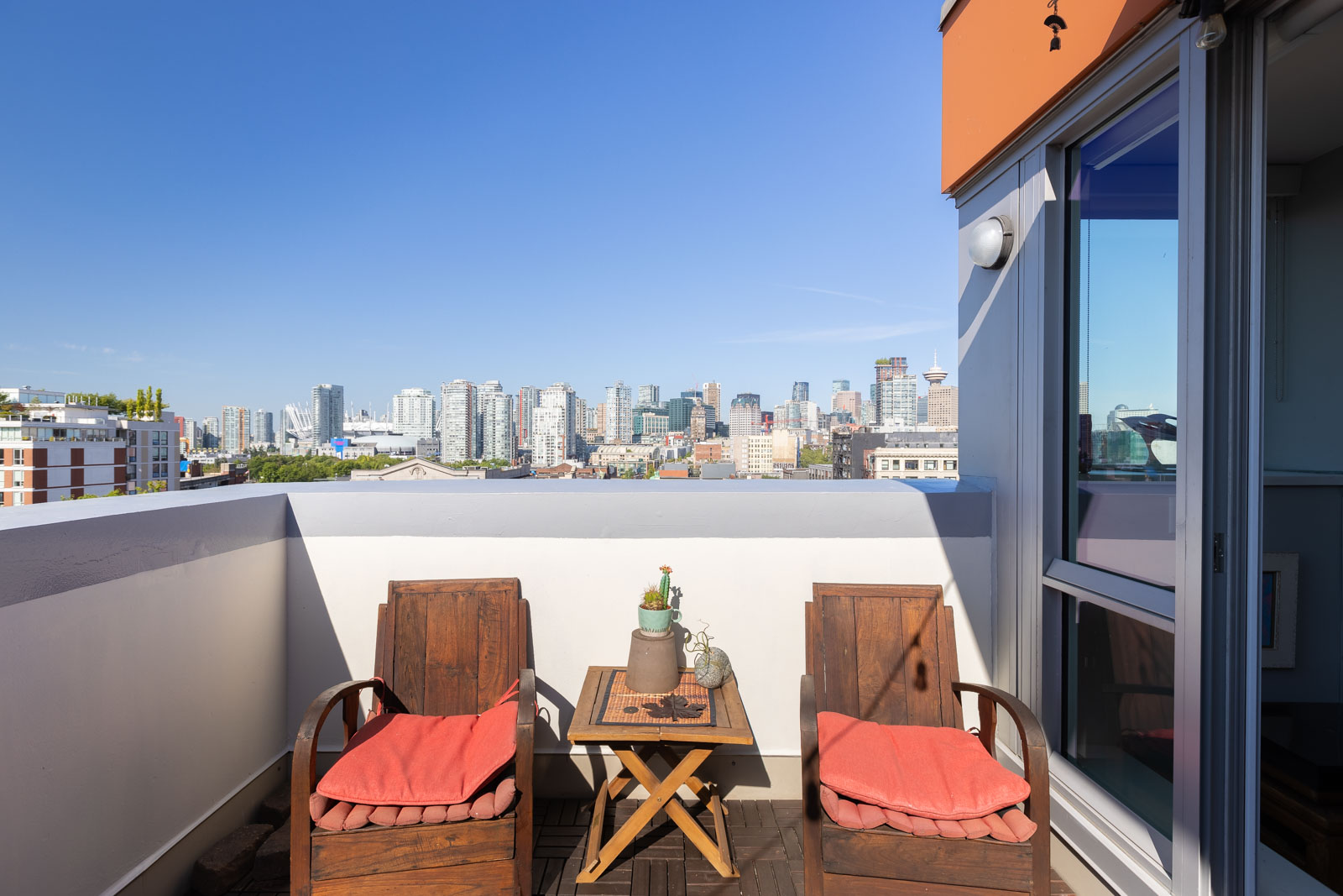 A city skyline view from a balcony with two wooden chairs, a table, and red cushions against a clear blue sky.