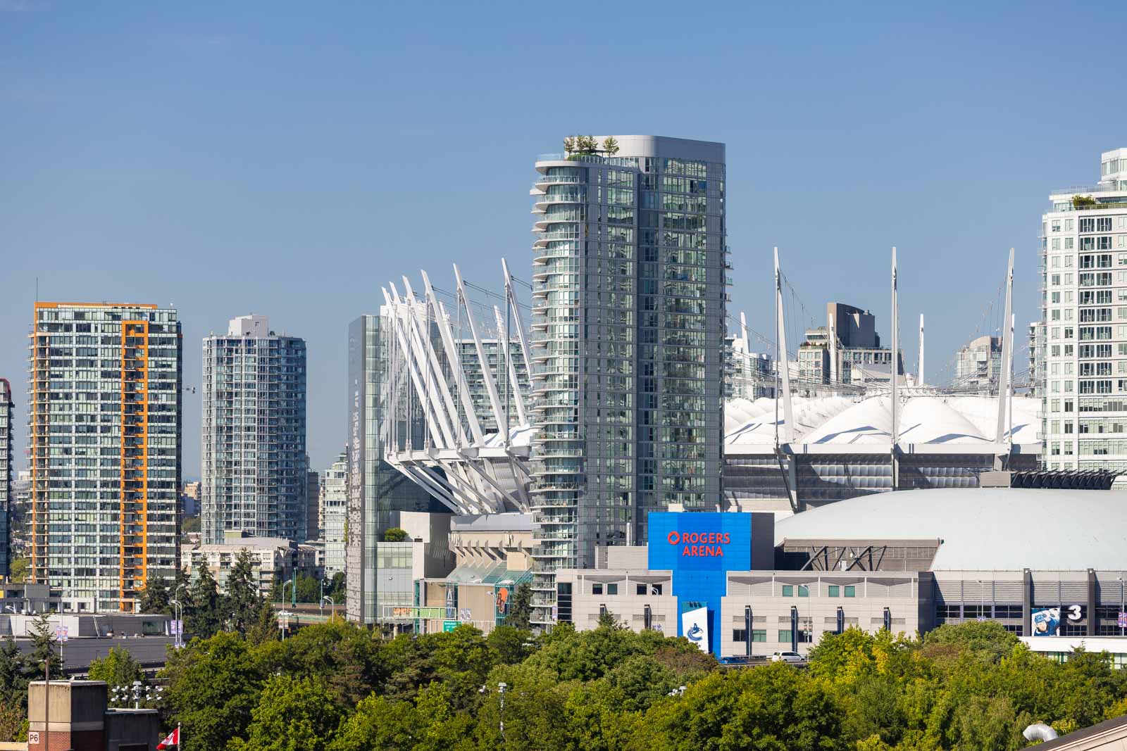 Vancouver skyline featuring skyscrapers with Rogers Arena and BC Place stadium in the foreground, surrounded by trees under a clear blue sky.