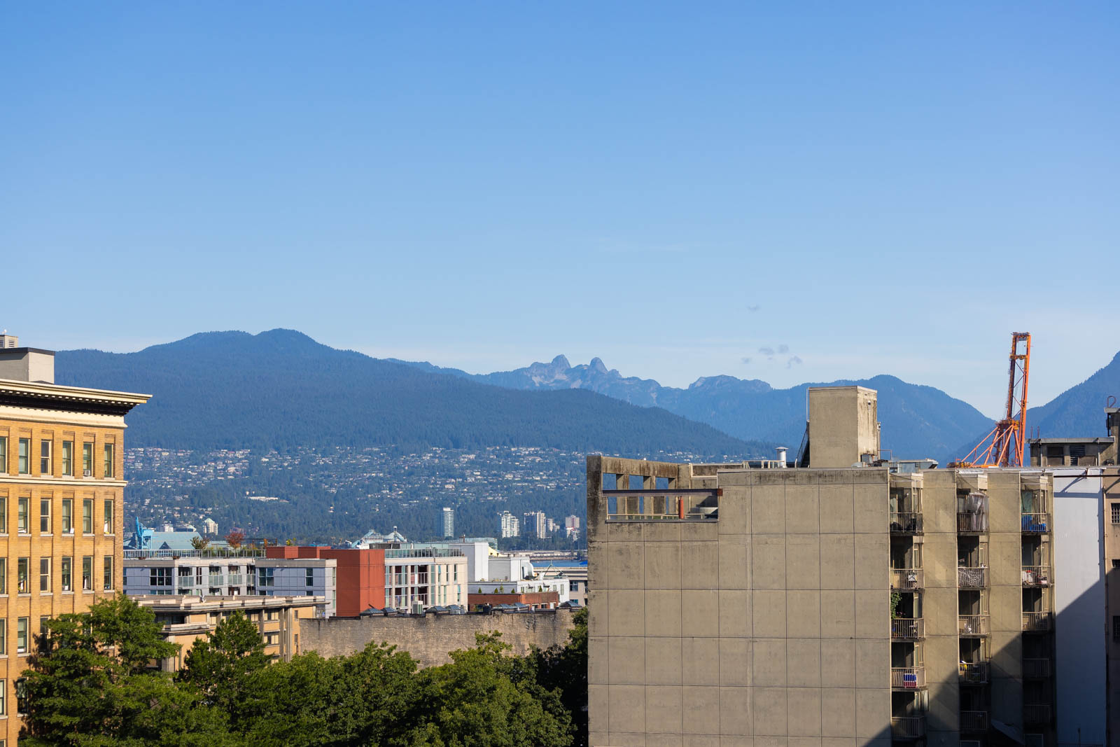 Cityscape with tall buildings in the foreground and tree-covered mountains under a clear blue sky in the background.