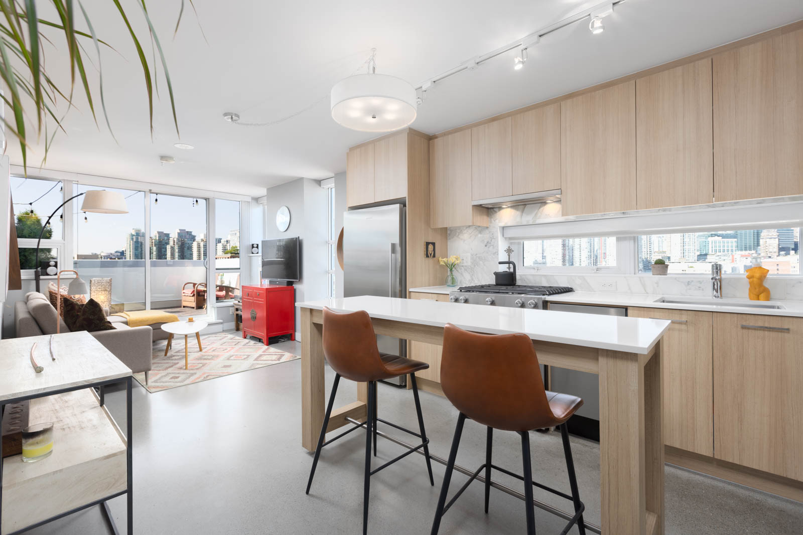 Modern kitchen and living area with light wood cabinets, stainless steel appliances, and an island with two brown chairs. Large windows offer a city view and natural light.