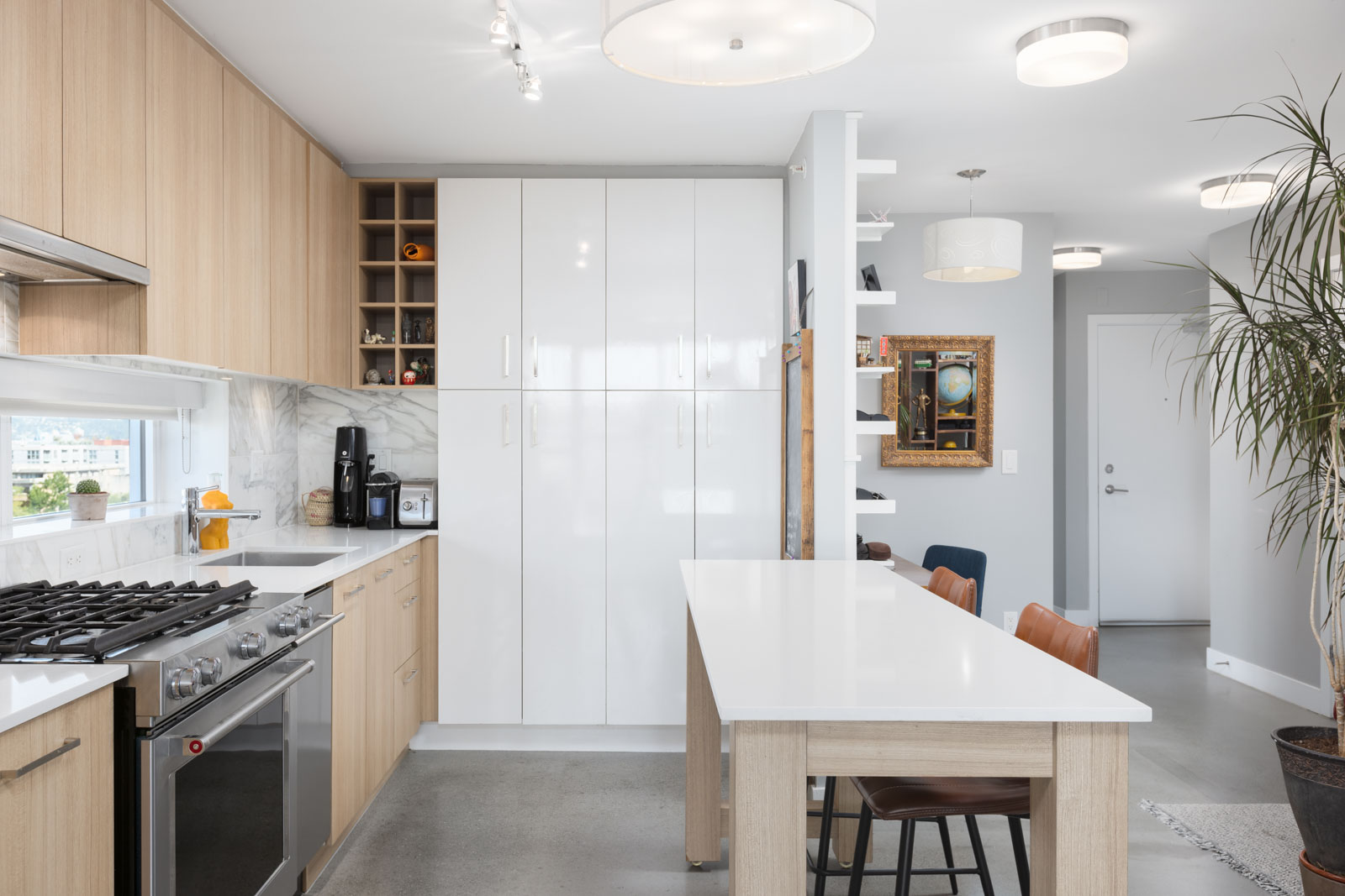 Modern kitchen with light wood cabinetry, white countertops, stainless steel appliances, and a small dining area with chairs. Large potted plant by the entrance, and decorative items on shelves.