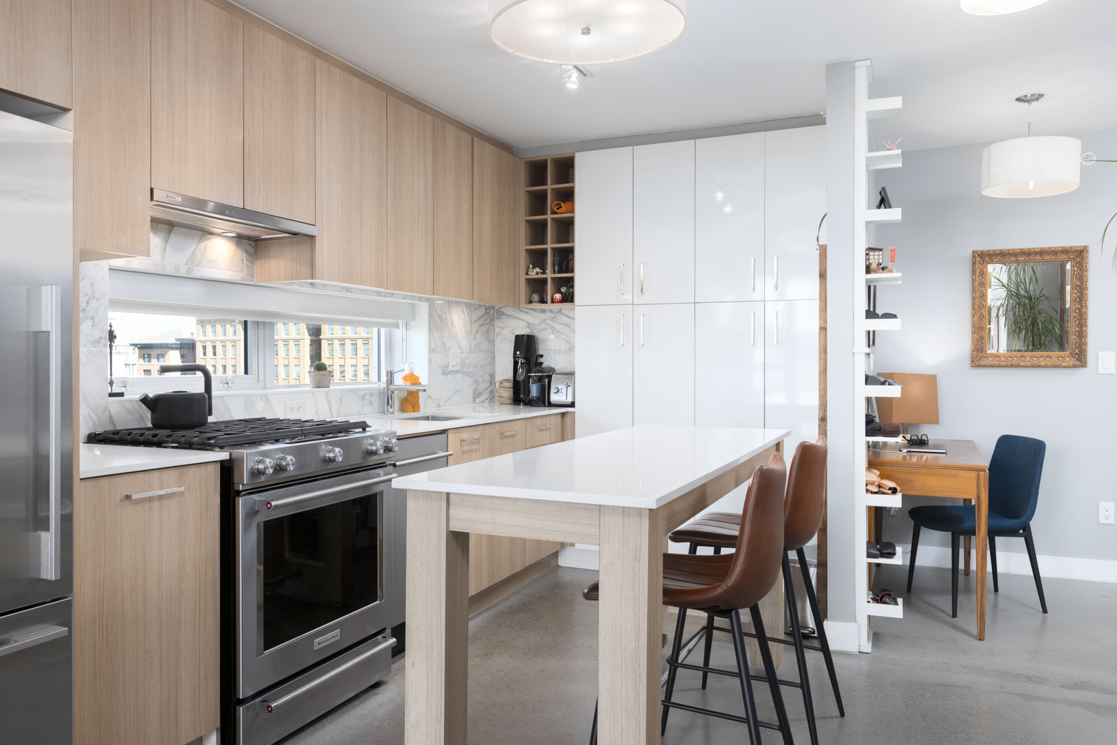 Modern kitchen with light wood cabinets, stainless steel appliances, and a white marble backsplash. An island with bar stools is in the center. A small desk area and bookshelf are in the background.