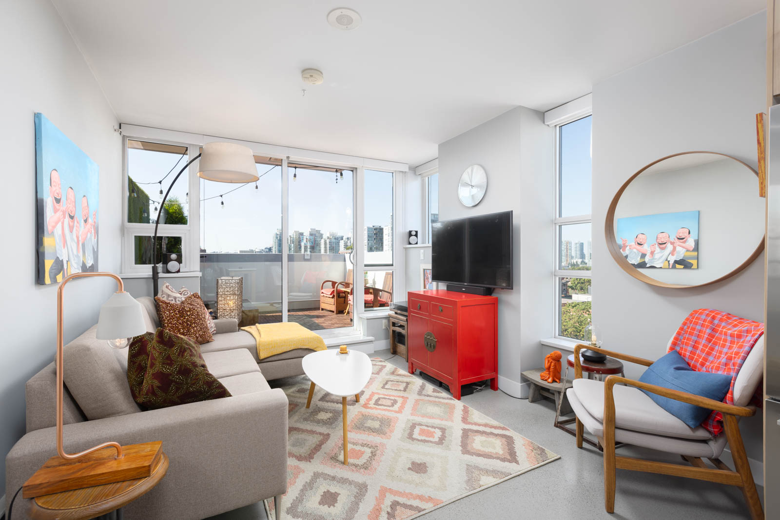 A modern living room with a gray sofa, yellow stool, red cabinet, and TV. Large windows provide city views. Decor includes a round mirror, abstract paintings, and a patterned rug.