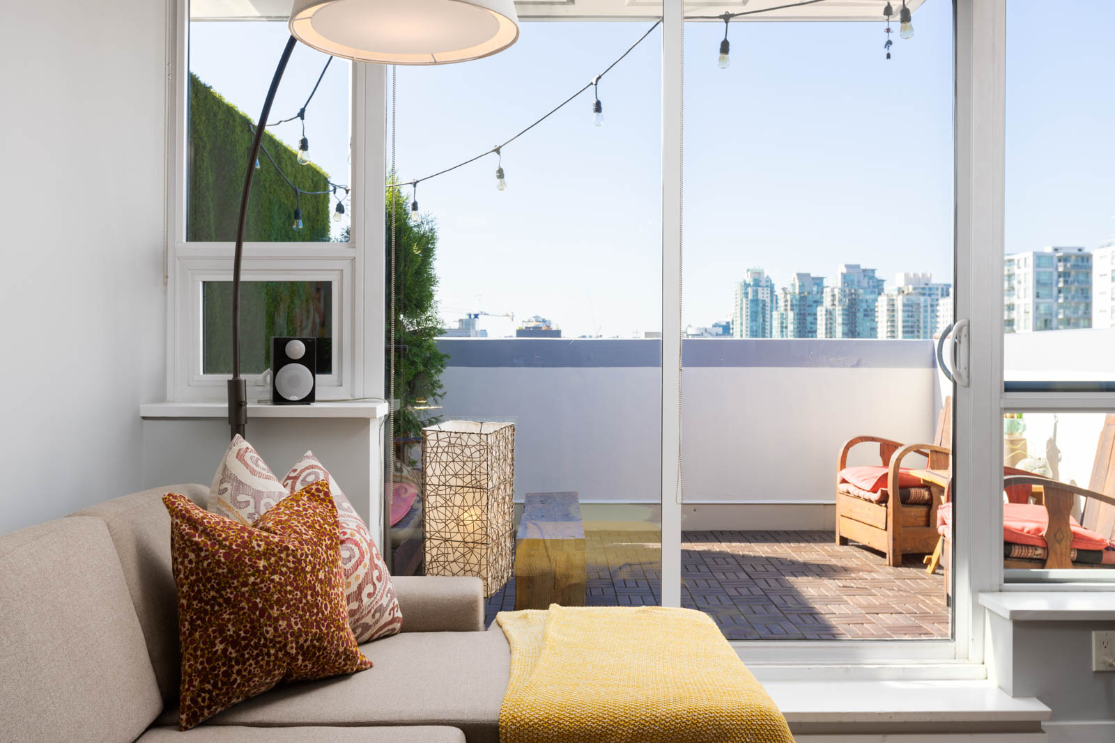 Living room with beige sofa, colorful cushions, and yellow throw. Floor lamp beside. Glass doors open to a balcony with seating and city skyline view. String lights hang outside.