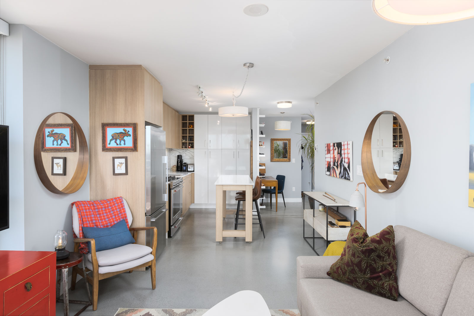 Modern open-plan living room and kitchen with neutral tones. Features include a beige sofa, armchair, round mirrors, and wall art. Natural light from large windows brightens the space.