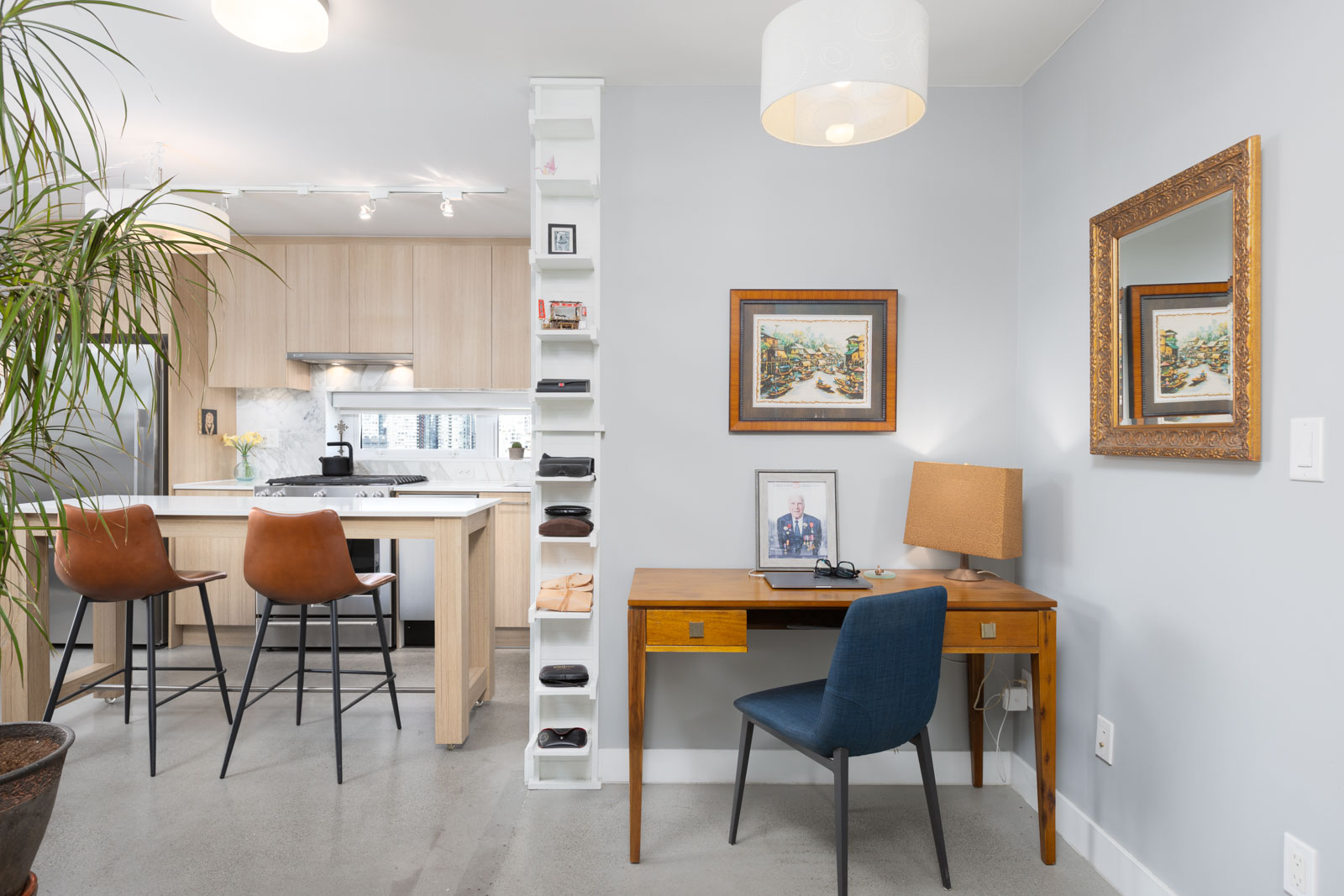 Home office and kitchen area with a wooden desk, blue chair, lamp, framed pictures, bar stools, and a kitchen island with plants and decor items.