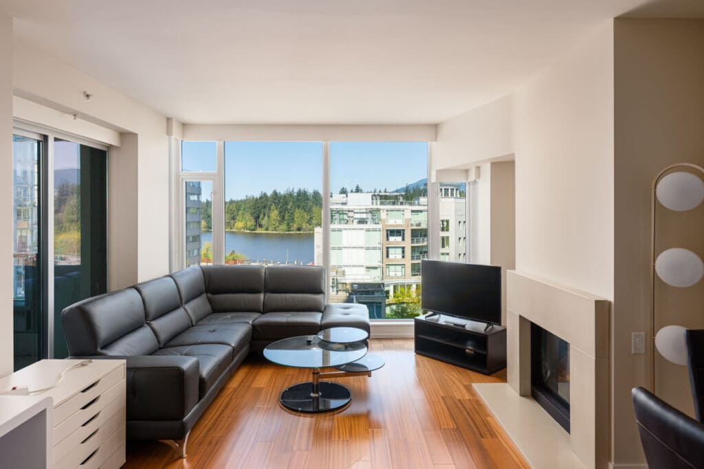 Modern living room with a black sectional sofa, glass coffee tables, TV, and large windows offering a view of city buildings, trees, and water.