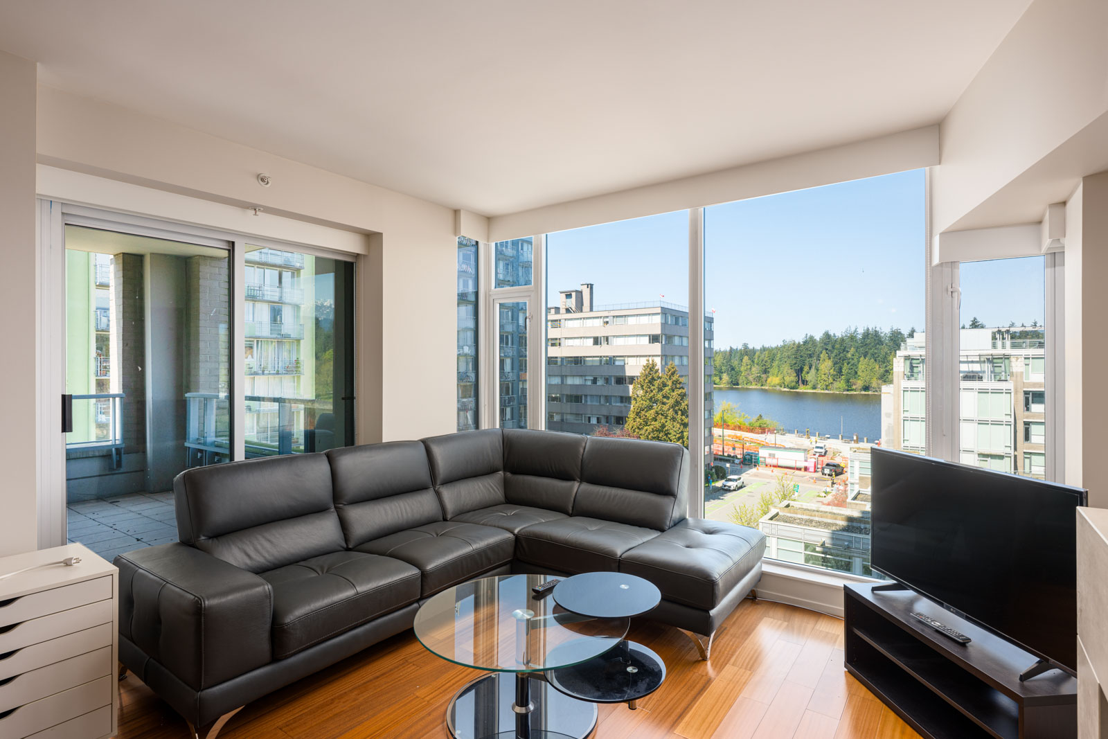 Modern living room with black sectional sofa, glass coffee table, TV, large windows, and balcony access, overlooking nearby buildings, trees, and a body of water.