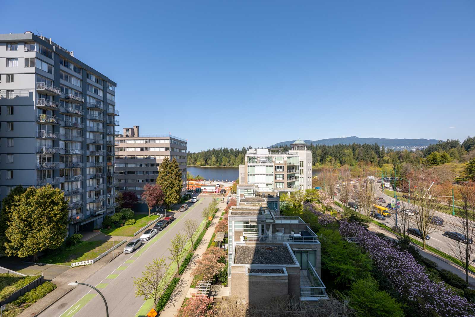View of a city street lined with mid-rise apartment buildings, trees, and cars, with a forested area and mountains in the background under a clear blue sky.