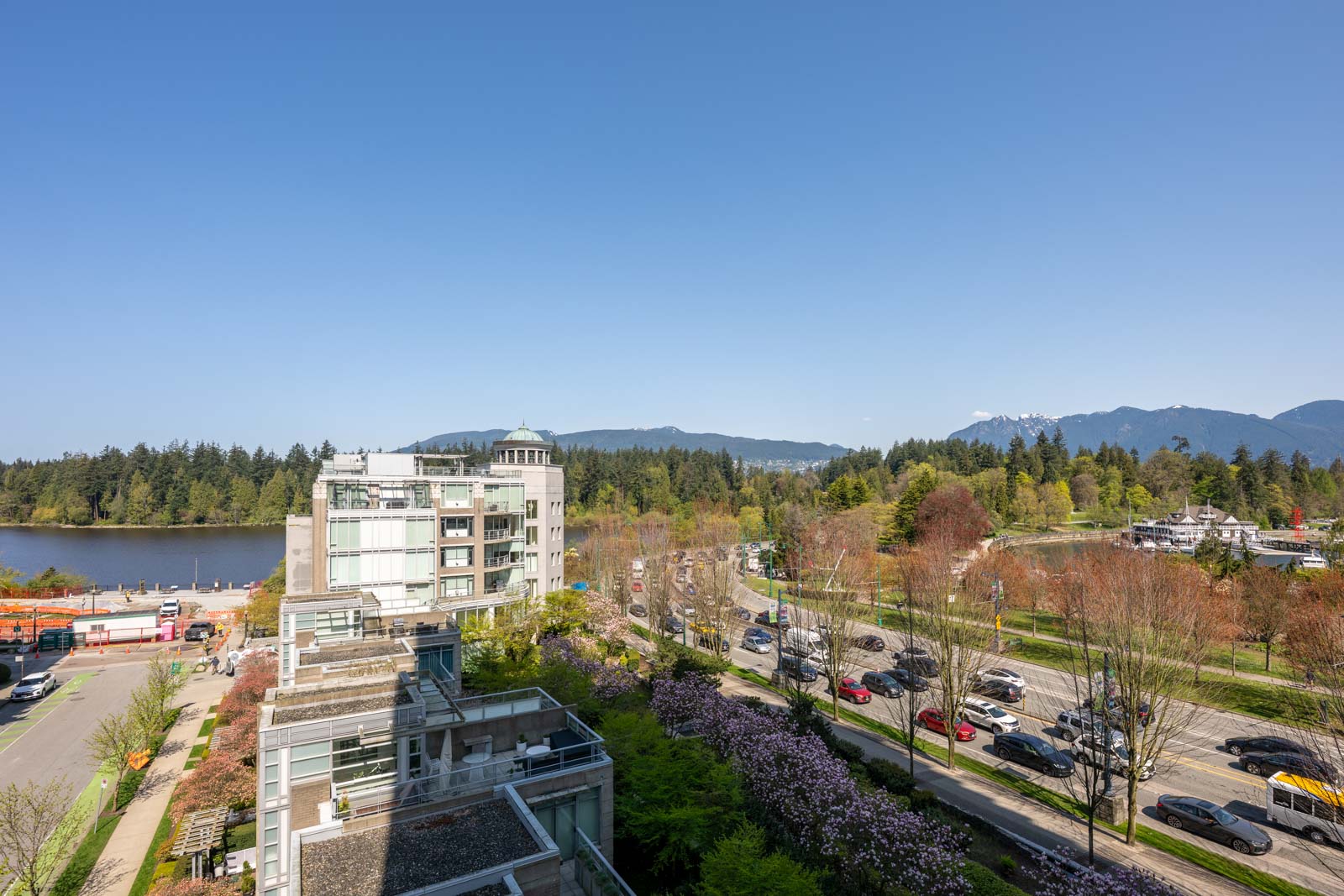 A city street with moving cars runs beside a park and glass buildings, with a lake, trees, and mountains in the background under a clear blue sky.
