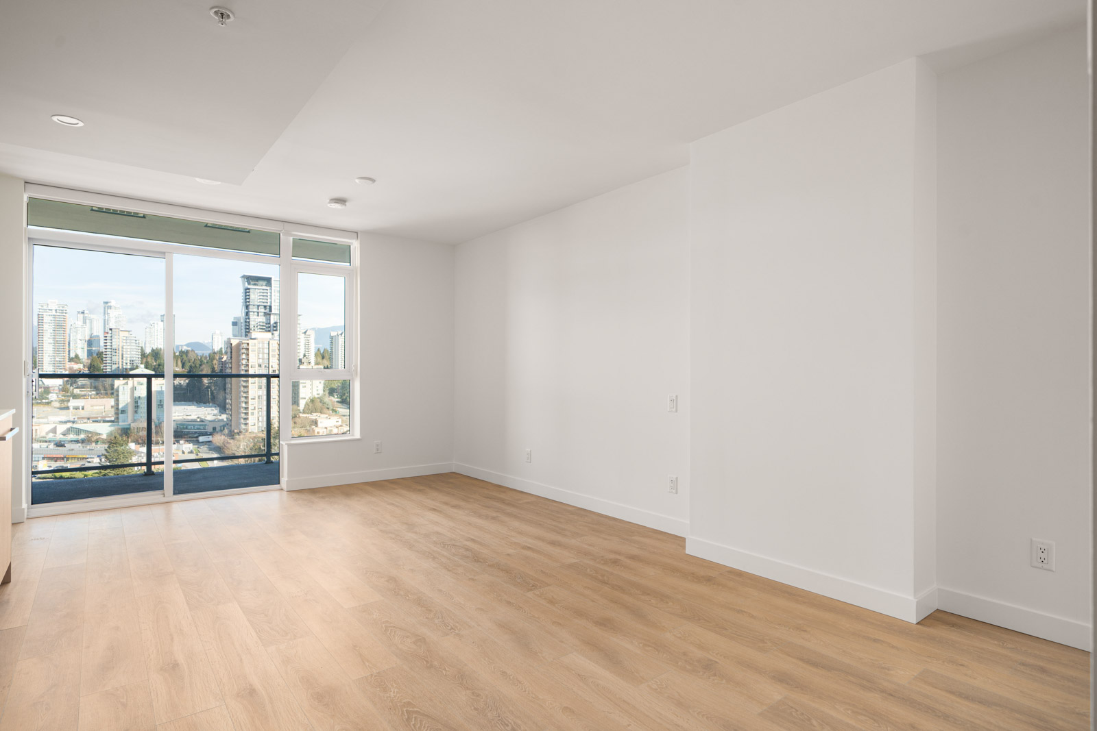 Empty, modern apartment with large window and balcony, wooden flooring, white walls, and ceiling lights, offering a view of city buildings in the distance.