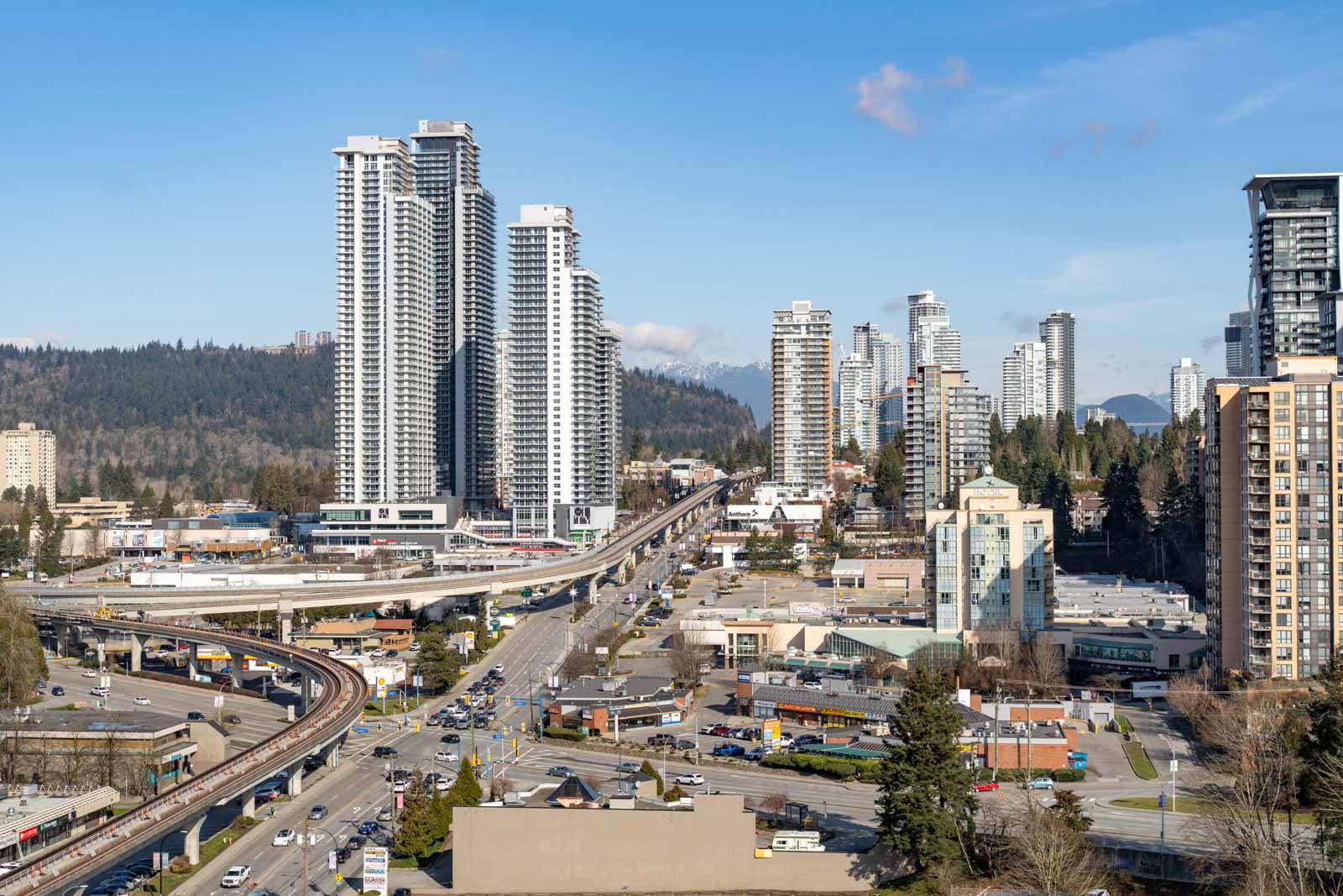 Aerial view of a cityscape with tall buildings, roads, and traffic, set against a backdrop of trees and mountains under a blue sky.