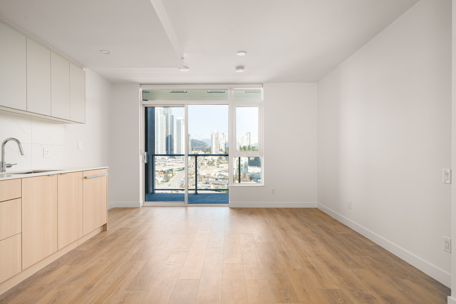 Empty modern apartment with wooden floors, light-colored cabinets, and large windows offering a city view.