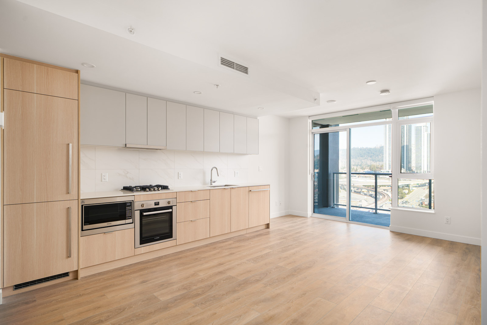 Modern, unfurnished kitchen and living space with light wood flooring, built-in appliances, and large windows leading to a balcony with a city view.