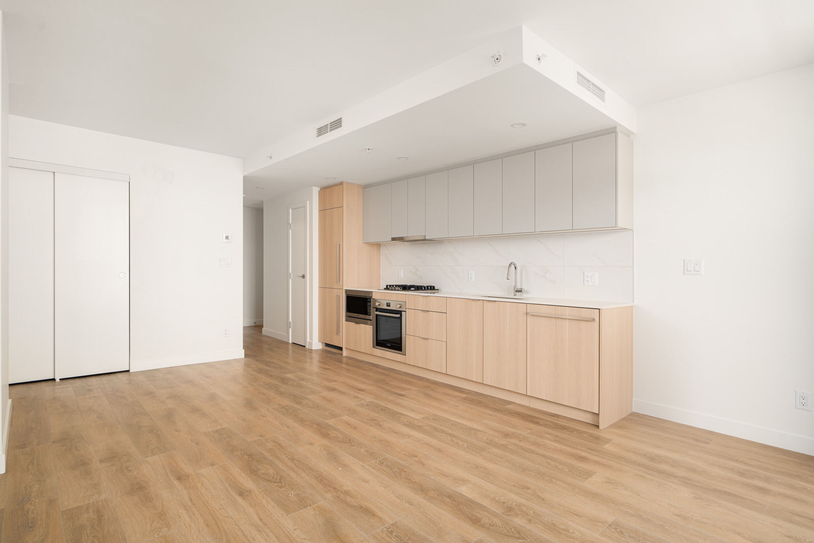 Minimalist kitchen with light wood cabinets, built-in oven, and sink on a white countertop. Open floor plan with wood flooring and white walls.