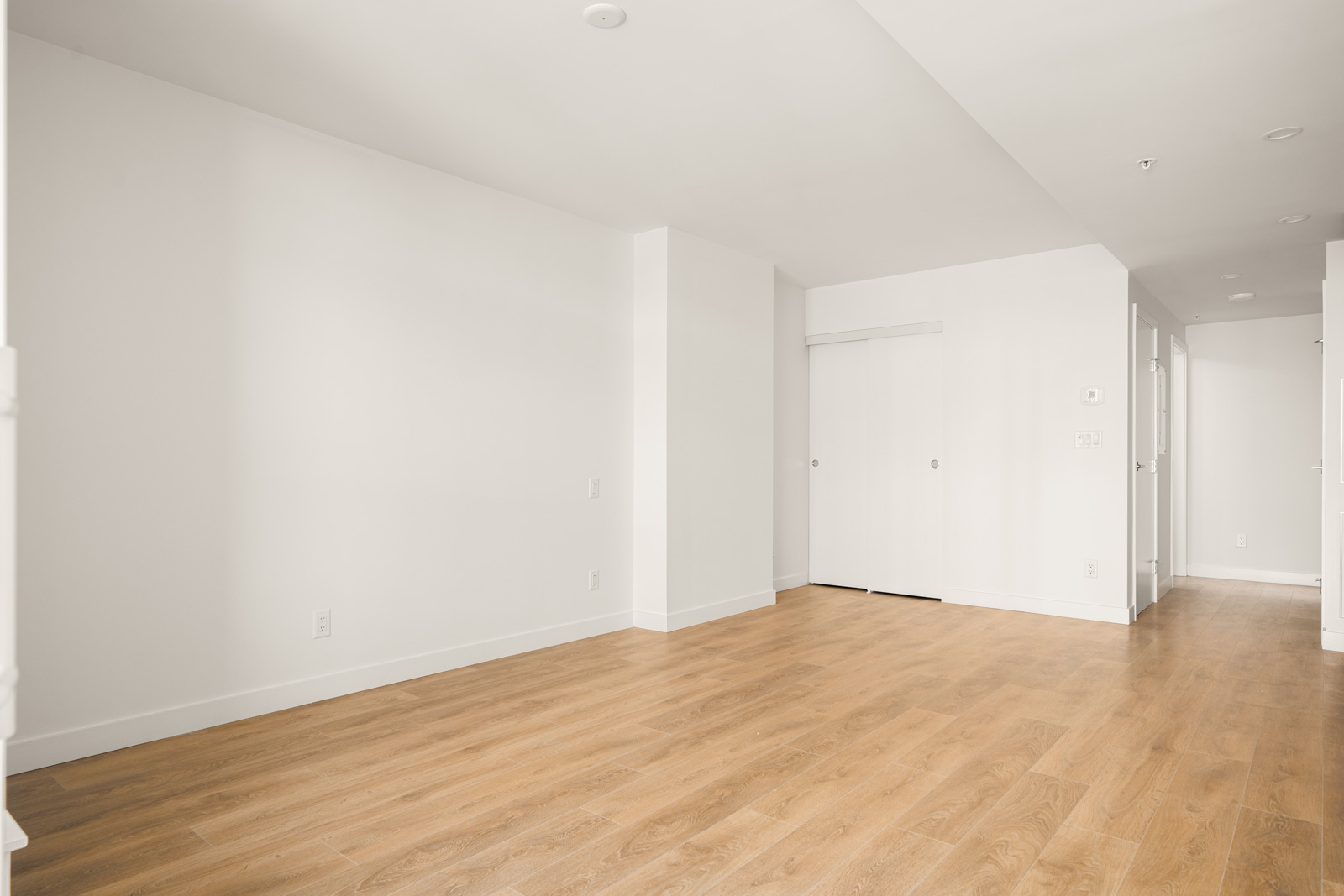 Empty room with light wooden floors, white walls, and a closed sliding door. Hallway visible to the right.