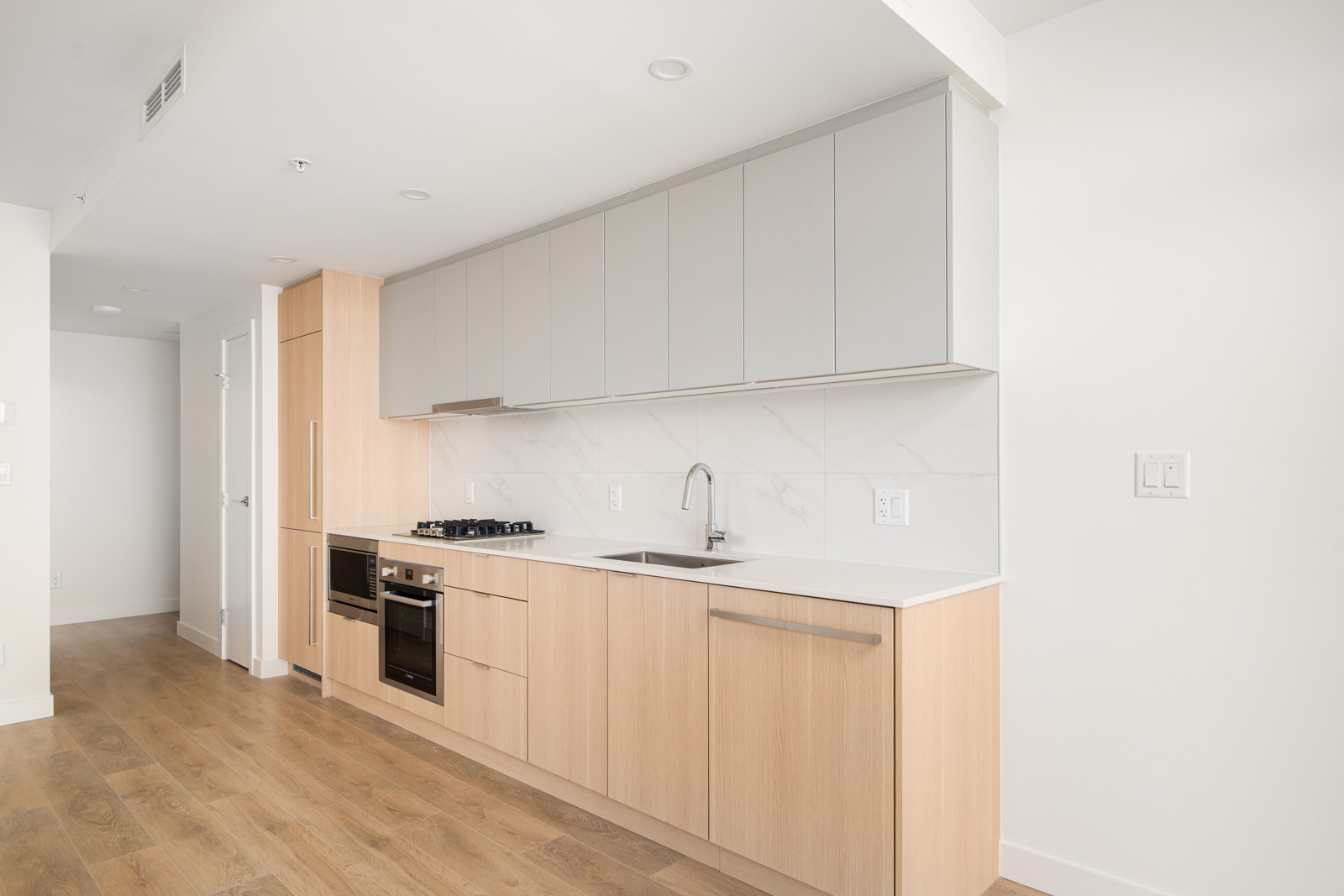 Modern kitchen with light wood cabinets, white countertops, gray upper cabinets, built-in oven, stove, and stainless steel sink on wooden flooring. White walls and minimal design.