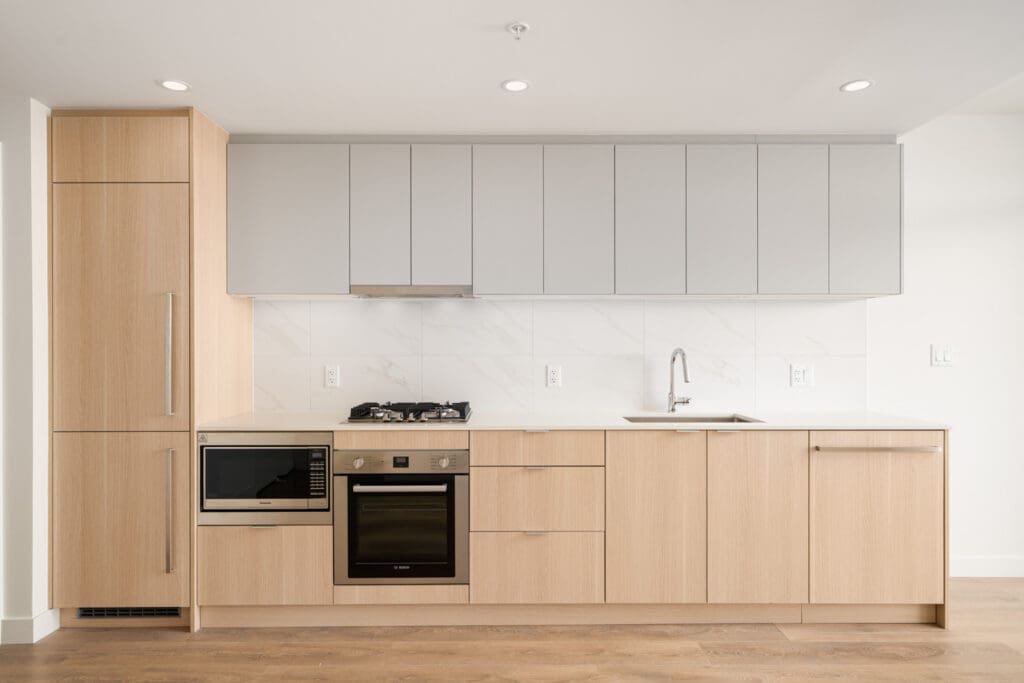 Modern kitchen with light wood cabinetry, white countertops, and backsplash. Features a built-in oven, microwave, gas stove, and sink. Simple, minimalist design.