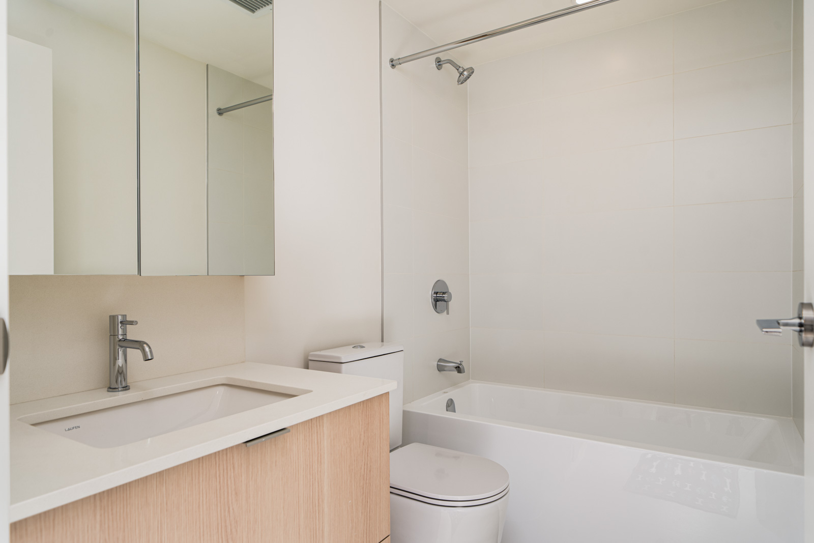 Modern bathroom with a white bathtub, toilet, sink, and a mirrored cabinet. Walls and floors are light-colored. There is a chrome faucet and showerhead.