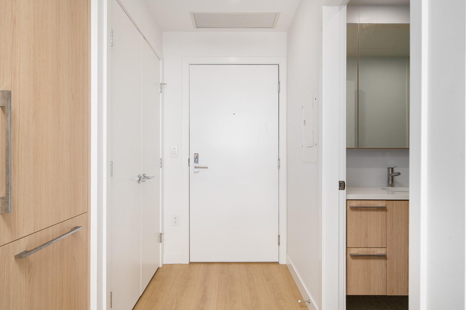 A hallway with light wood flooring, a white front door, and built-in wooden cabinets. A partially open door reveals a bathroom with a small sink and mirror.