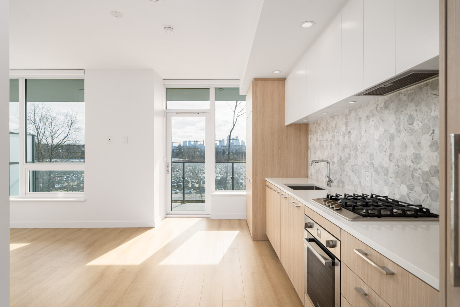 Modern kitchen with light wood floors, white cabinets, hexagonal tile backsplash, built-in stove and oven. Large window and glass door leading to a balcony with city view.