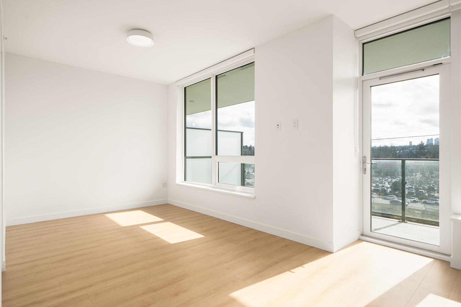 A bright, empty room with wooden floors, white walls, large windows, and a glass door leading to a balcony with a city view.
