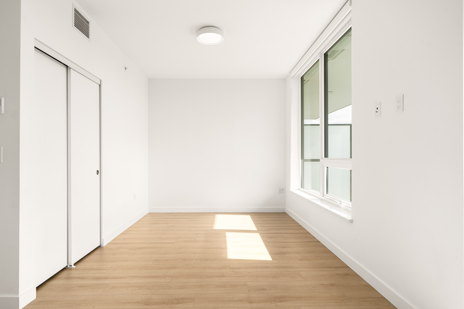 Empty room with light wooden floor, white walls, and a large window with a view. A closed closet with sliding doors is on the left. Ceiling light fixture is centered.