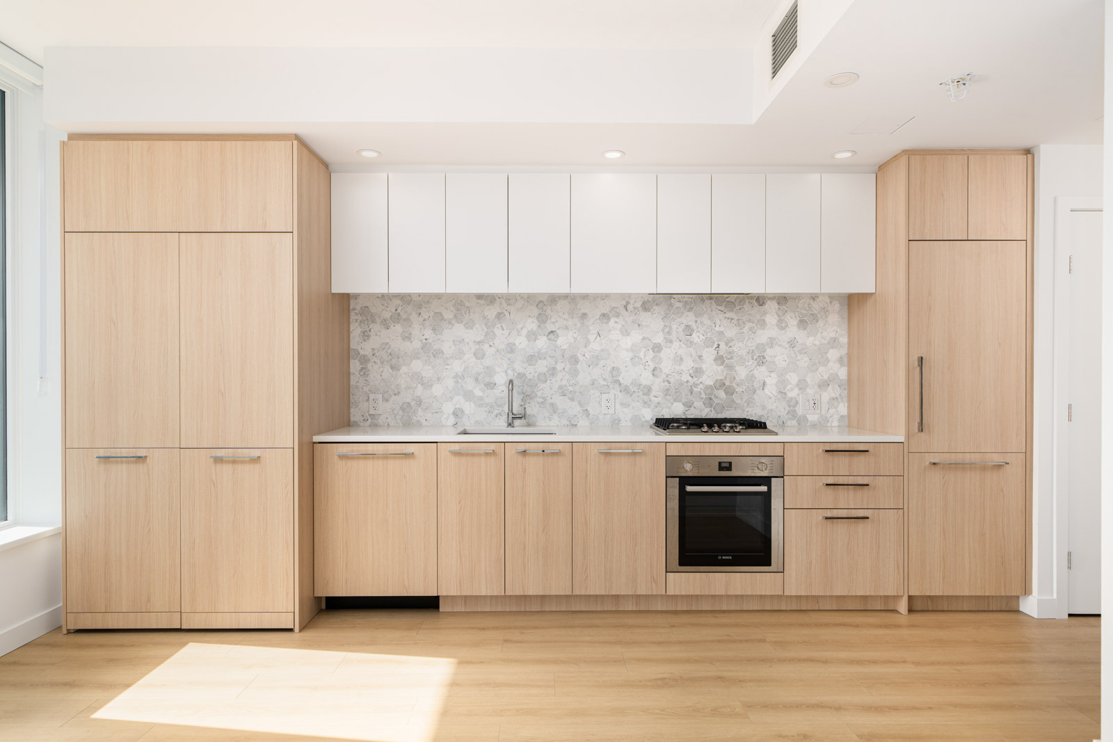 Modern kitchen with wooden cabinets, white upper cabinets, a floral-patterned backsplash, a built-in oven, and a small sink under bright lighting. Light wood flooring.