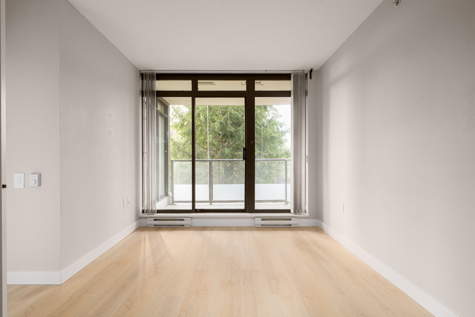 Empty room with light wood flooring, white walls, and a large windowed door leading to a balcony with a view of trees.