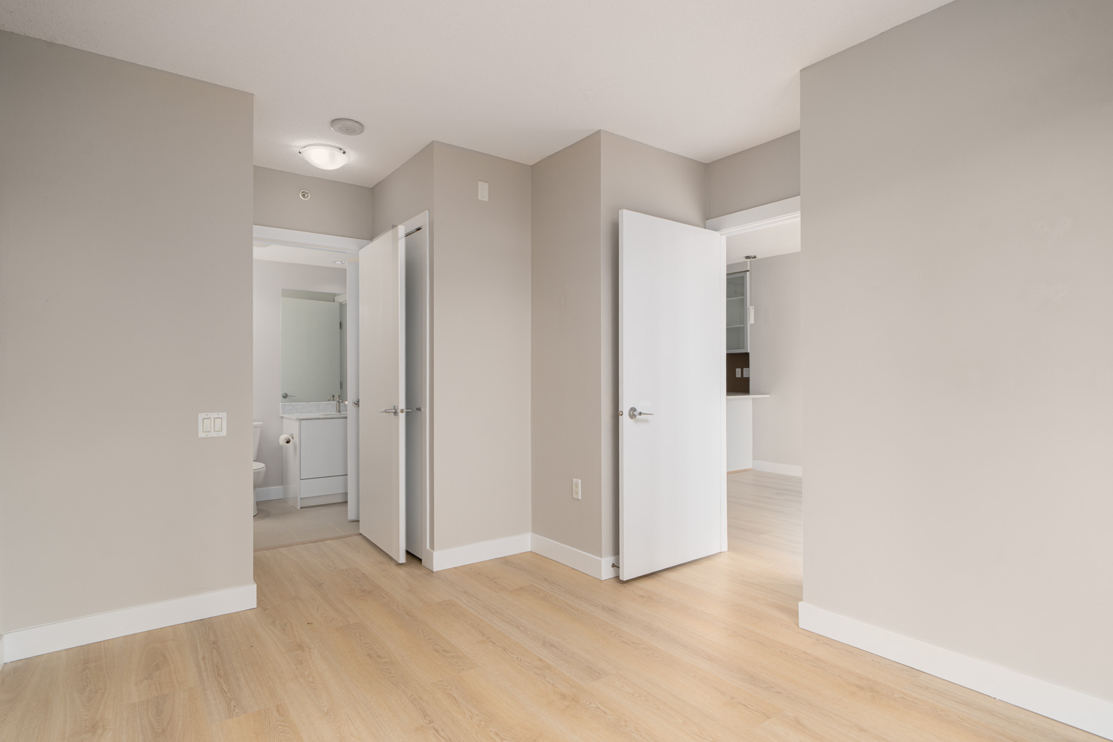 Empty room with light wood flooring, neutral walls, and two open doors leading to a bathroom and another room. Ceiling light fixture is visible.