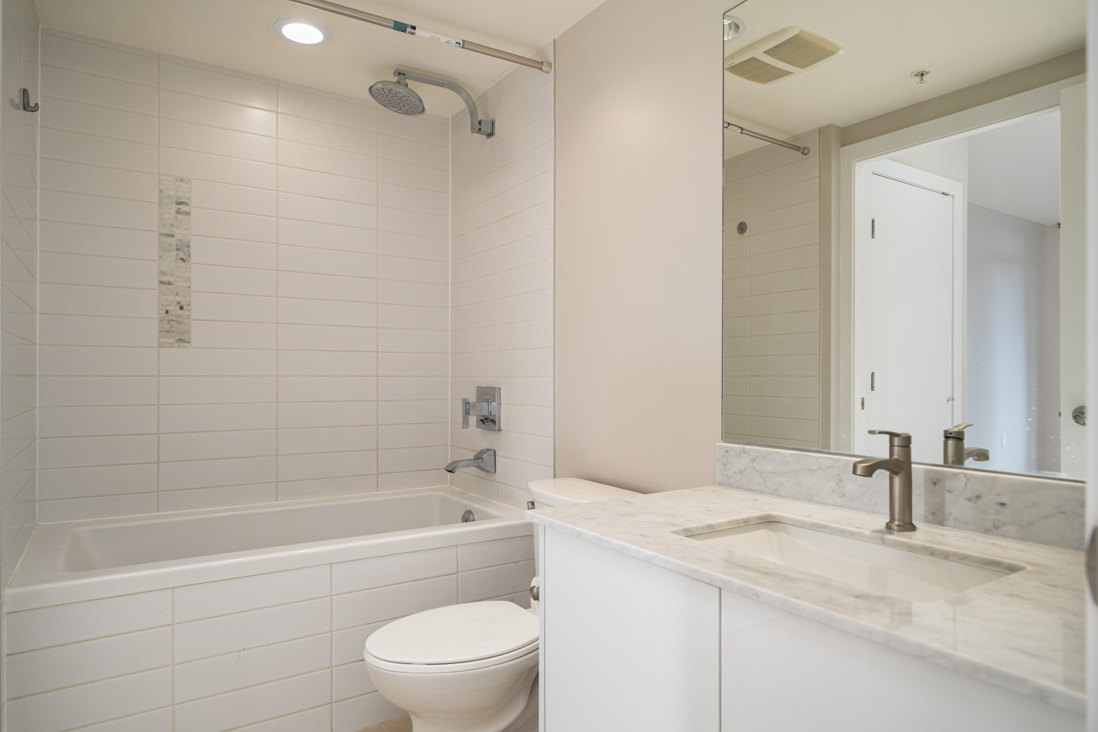 A modern bathroom with a white bathtub, overhead shower, toilet, and a countertop sink. The walls are tiled, and a mirror hangs above the sink.