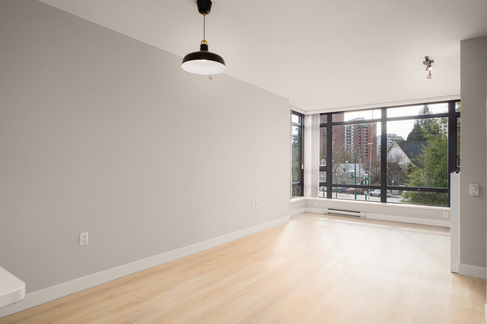 Empty room with light wood flooring, a large window showing trees and buildings, a black pendant light, and gray walls.