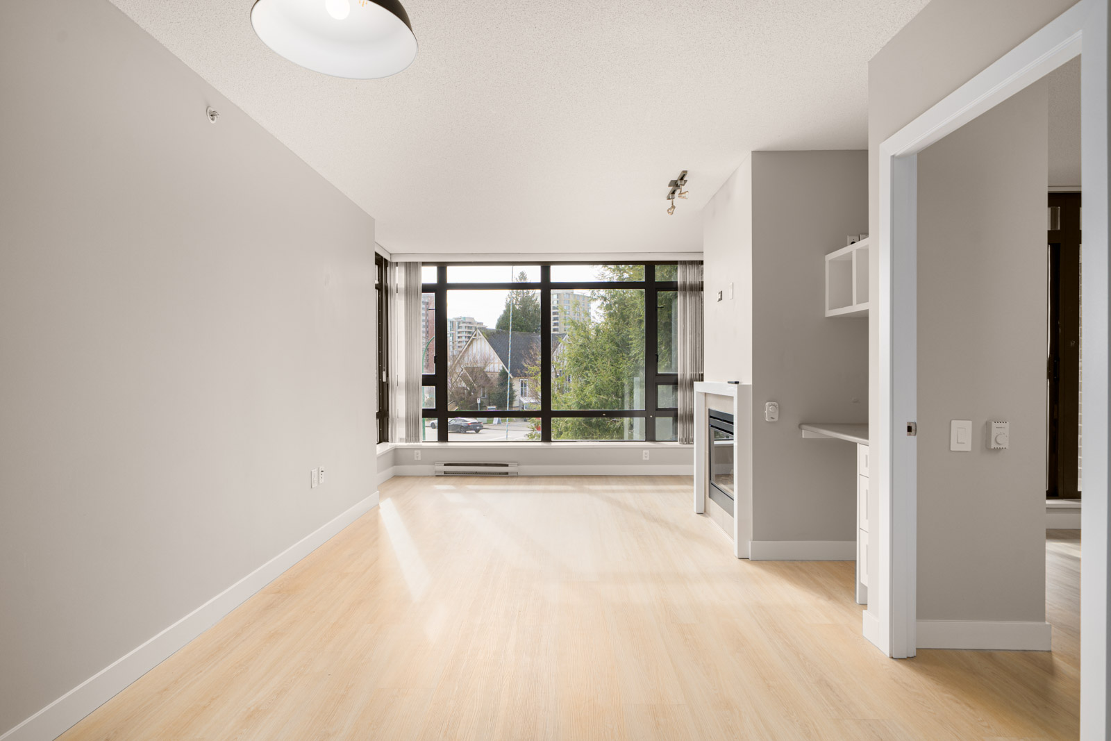Bright, empty apartment with light wood floors, large windows, and white walls. Includes a small built-in desk and shelves, with a view of trees and buildings outside.