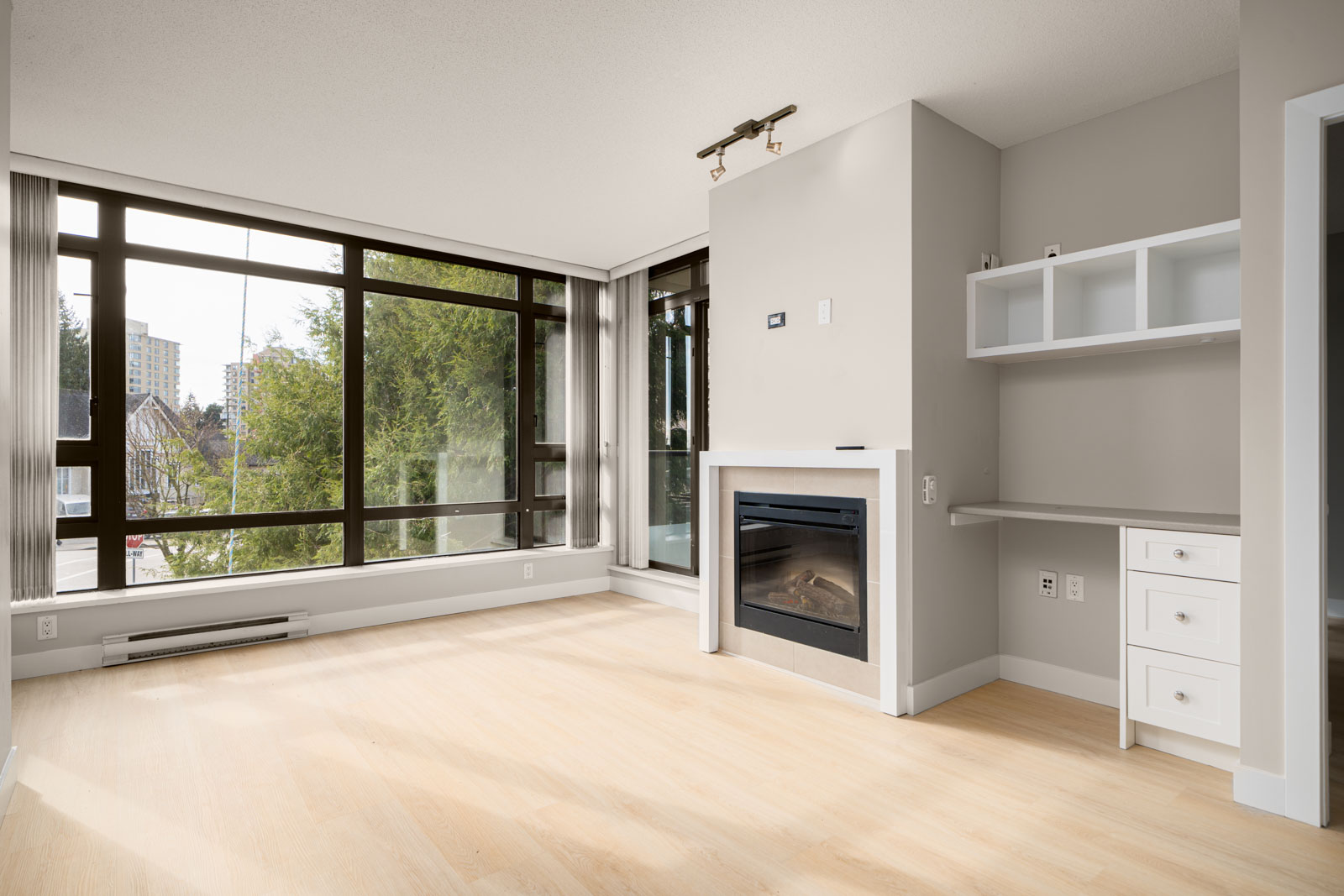 Modern, unfurnished living room with large windows, light wood flooring, a built-in desk with shelves, and a fireplace.