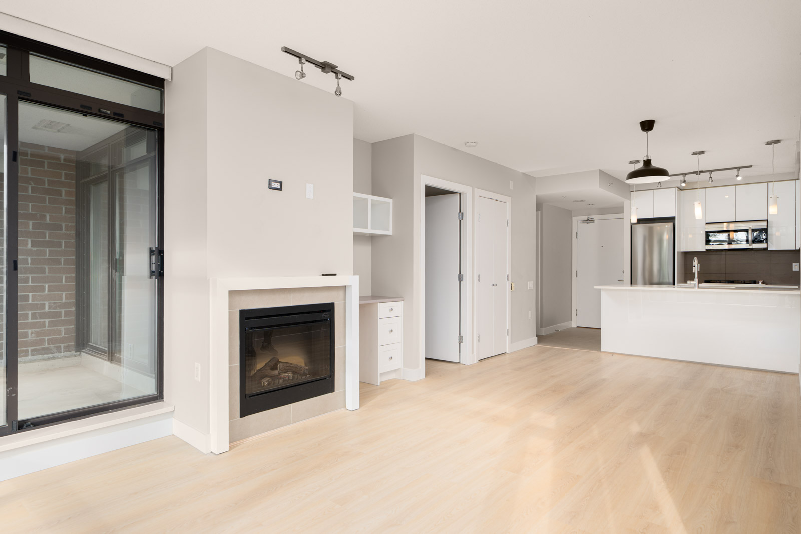 Modern, unfurnished apartment interior with light hardwood floors, a fireplace, large window, and a kitchen with white cabinets and stainless steel appliances.