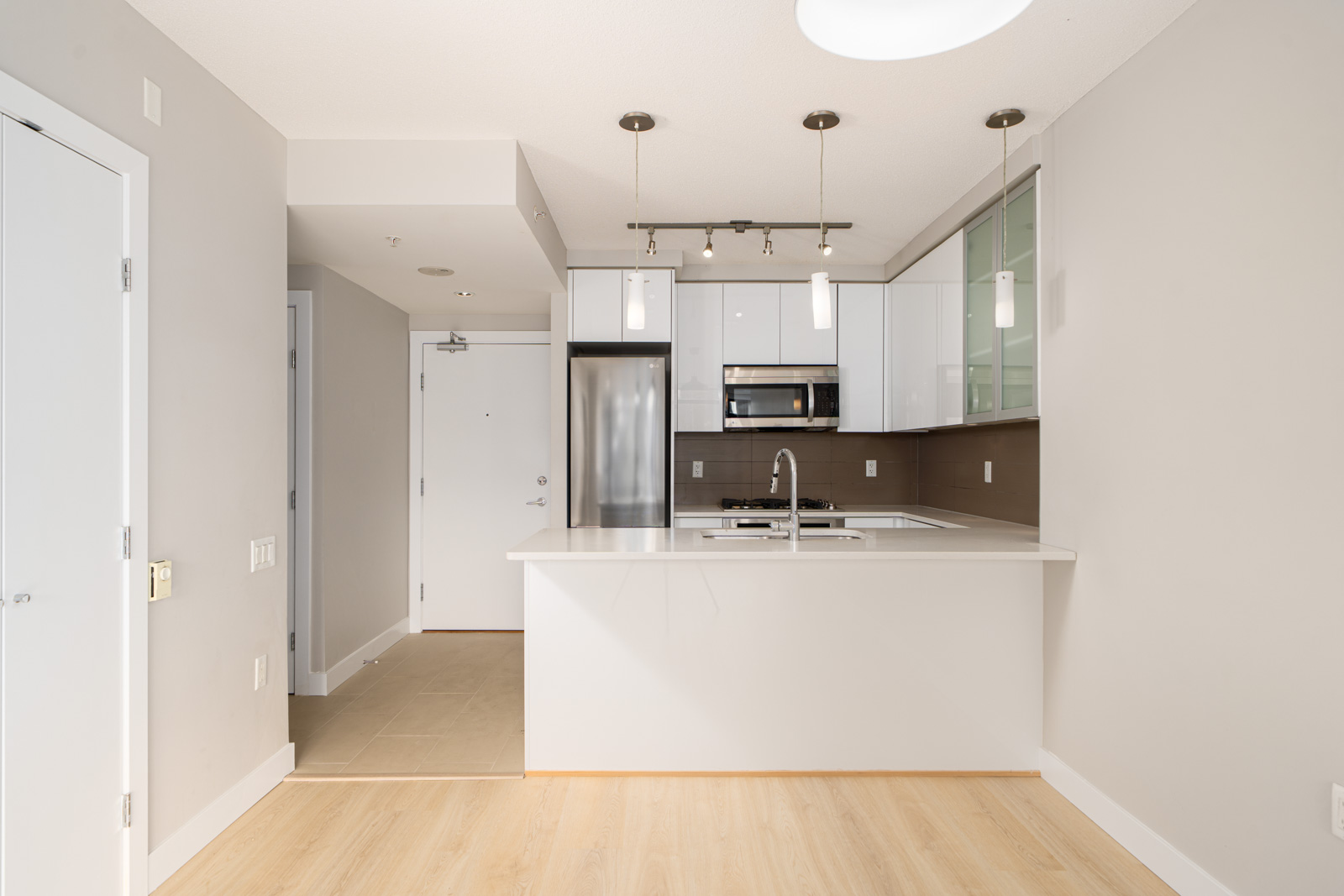Modern kitchen with white cabinetry, stainless steel appliances, and a peninsula counter. Wood flooring contrasts with the neutral walls. Pendant lights hang above the countertop.
