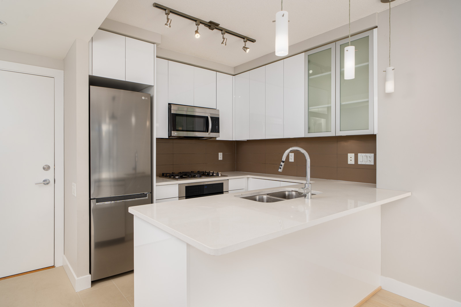 Modern kitchen with white cabinets, stainless steel appliances, a brown backsplash, and a white countertop island with a sink. Pendant and track lighting are visible above.