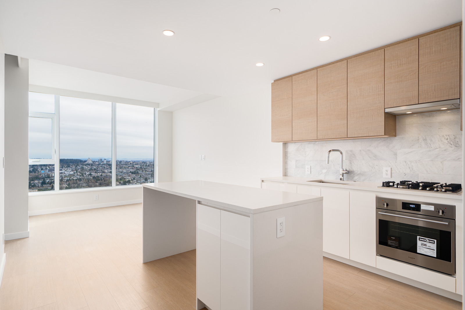 Modern kitchen with white cabinets, marble backsplash, and built-in appliances. An island with an extended countertop faces a large window overlooking a cityscape.