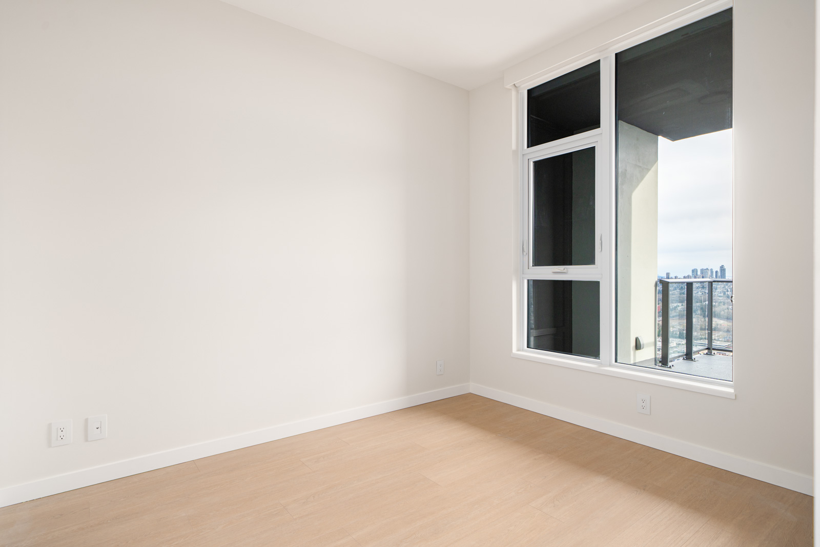 Empty room with light wooden floor, a large window showing a cityscape, and plain white walls.