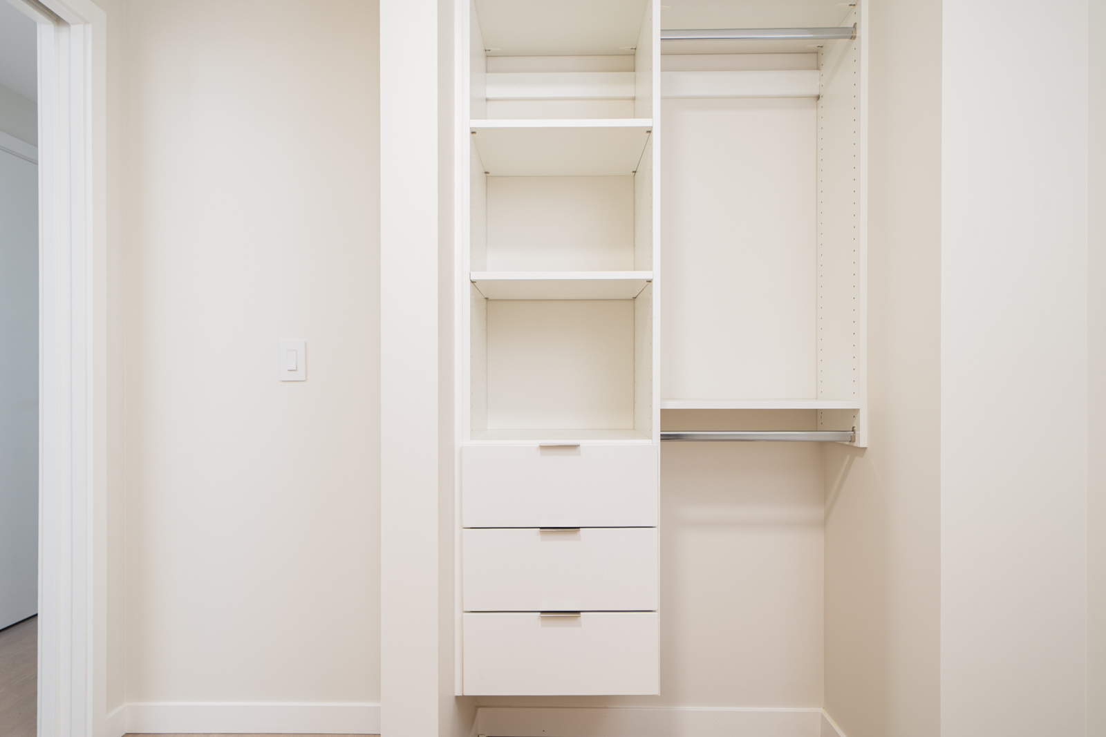 A white built-in closet with three shelves and three drawers on the left side, and a vertical hanging rod on the right side, set against a light-colored wall.
