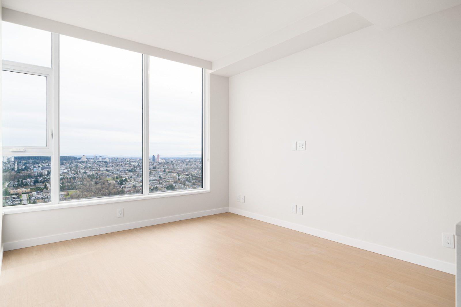Empty room with light wood flooring and large windows offering a cityscape view. Walls are white, and the room is well-lit.