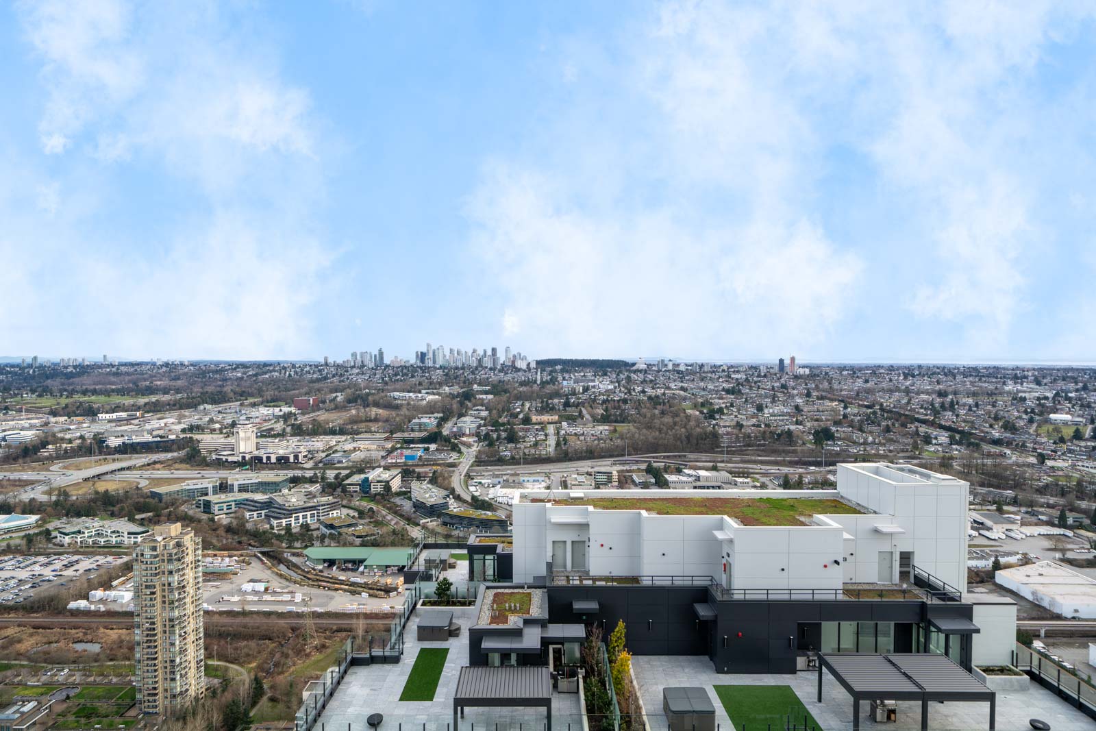 Aerial view of a cityscape featuring a modern building in the foreground with green rooftops, sprawling urban areas, and a distant skyline under a partly cloudy sky.