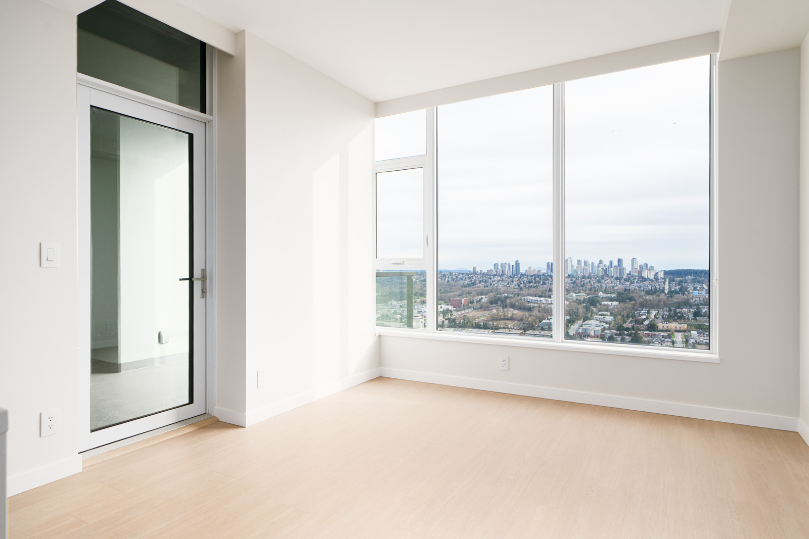 Empty room with light wood flooring, white walls, and large windows offering a city skyline view. A glass door is on the left side of the room.