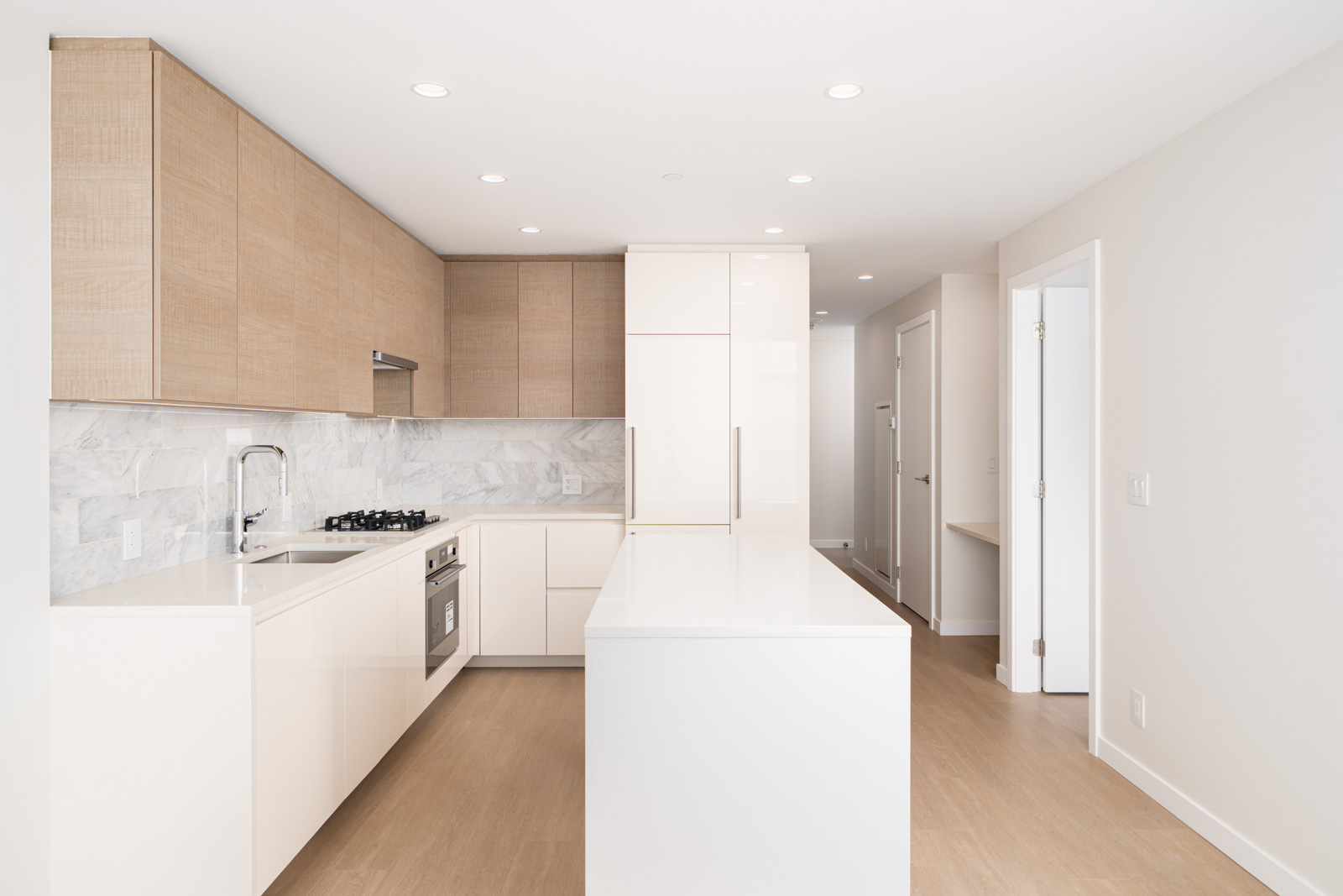 Modern kitchen with marble backsplash, white cabinets, wood accents, island, and recessed lighting. Stainless steel appliances are integrated into the design. Light wooden flooring throughout.