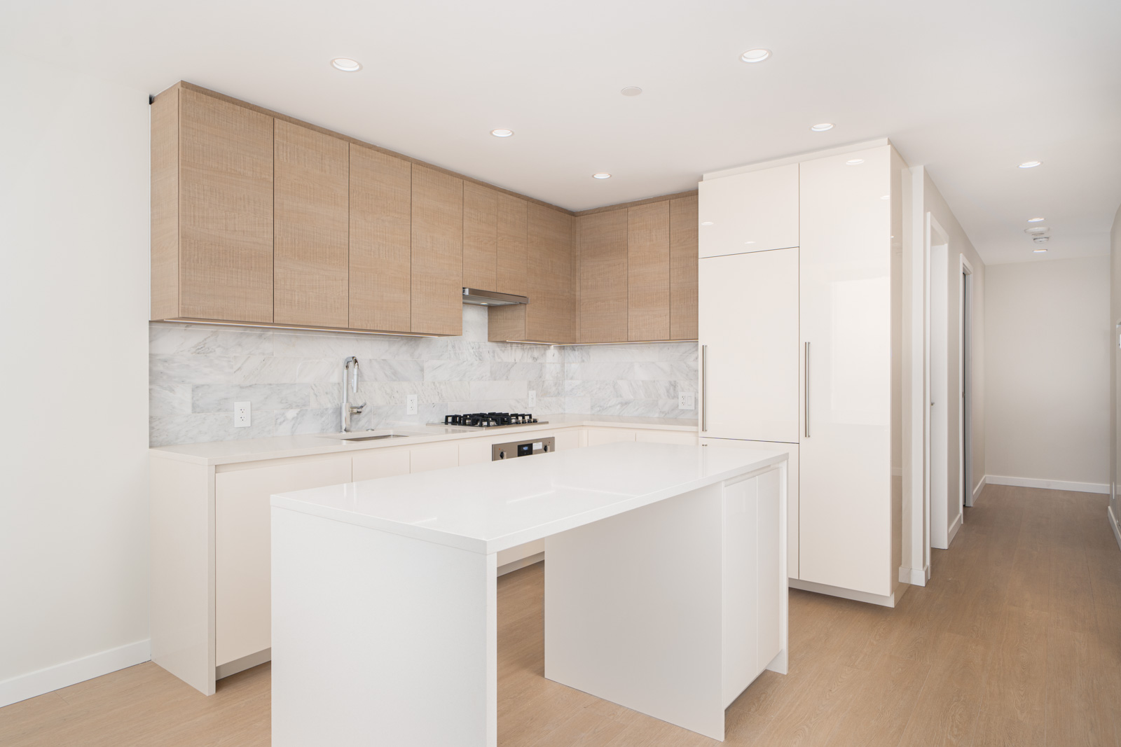 Modern kitchen with white countertops, light wood cabinets, and an island. Marble backsplash, built-in stove, integrated white refrigerator. Recessed lighting and wood floors.