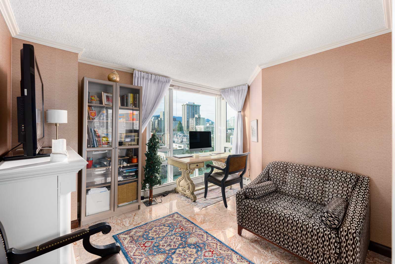 A small home office with a patterned sofa, desk, computer, bookshelf, and large window overlooking a cityscape.