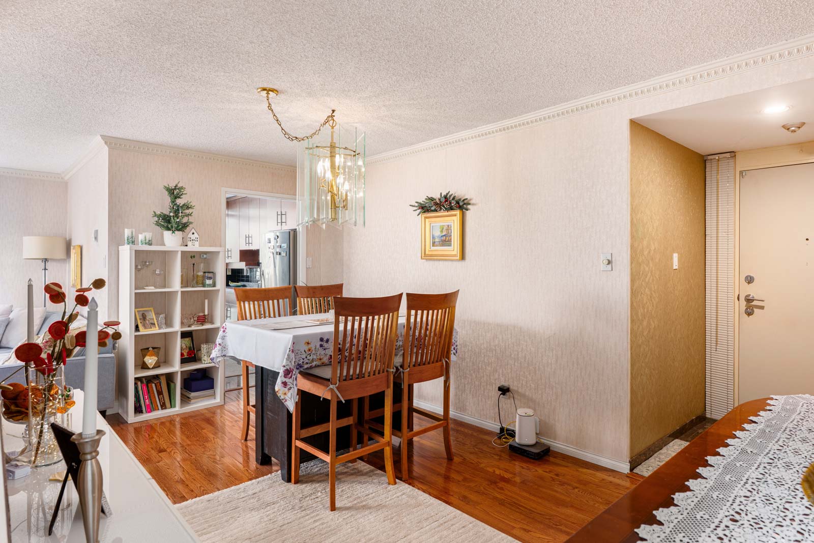 Dining area with a wooden table and chairs, floral tablecloth, chandelier above, bookshelf in the background, and a decorated wall. Doorway and entryway visible on the right.